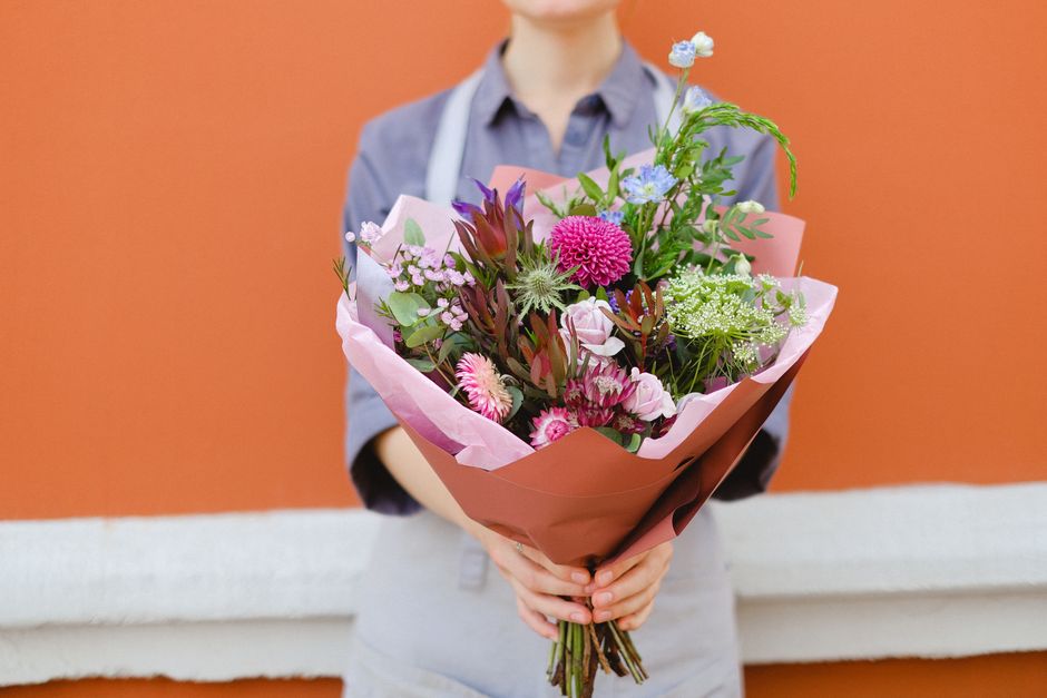 A person in a grey apron holds a vibrant bouquet arranged with a variety of fresh flowers including pink and purple blooms, green foliage, and delicate blue accents, wrapped in light pink and brown pa