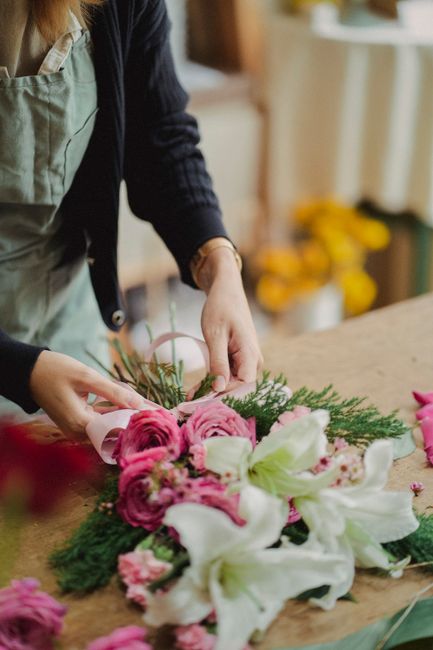 A professional florist from floristmerton.co.uk is arranging a floral gift on a wooden work surface, featuring a bouquet of fresh pink roses, white lilies, and green foliage. The florist is wearing a 
