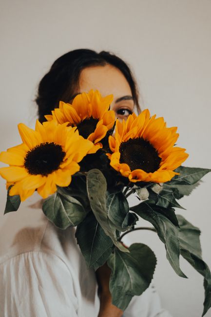 A woman with dark hair partially obscured by a large bouquet of vibrant yellow sunflowers with dark centers and broad green leaves, held close to her face. The flowers are fresh, with slightly texture