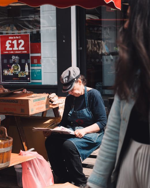 A woman sitting on a wooden bench outside a shop, reading a menu or brochure, wearing a grey hat, black shirt, and blue apron. In front of her, there are boxes of bread labeled 'Stronghold' and other 