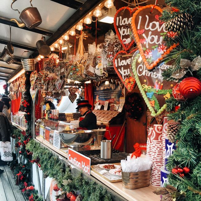 A festive market stall decorated with Christmas greenery and red accents, displaying a variety of gingerbread cookies featuring decorated heart-shaped designs with icing messages such as 'I love you' 