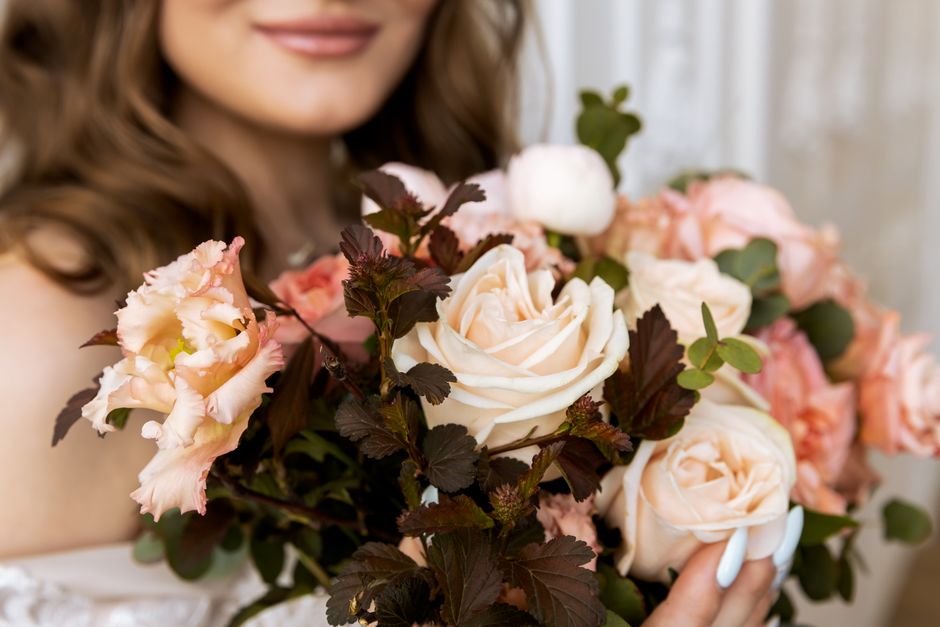 A close-up of a person holding a floral arrangement featuring large, cream-colored roses and soft pink lisianthus flowers, accented with dark green and burgundy foliage. The bouquet is arranged in a n