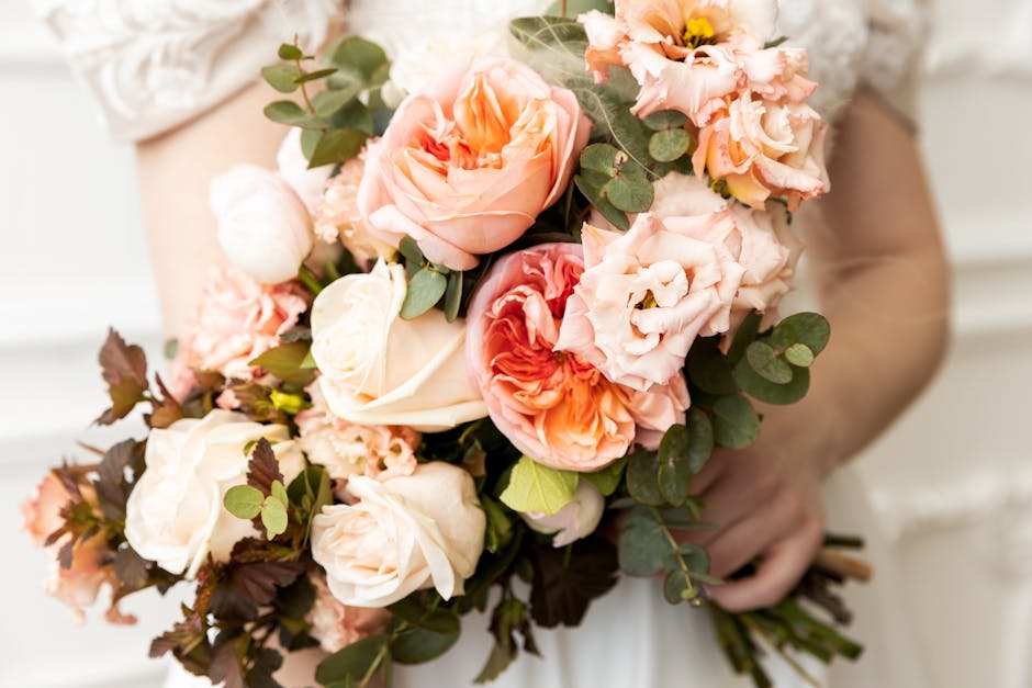 A person holding a bouquet of fresh flowers featuring large pink roses, peach lisianthus, and pink carnations, accented with eucalyptus leaves and dark-stemmed foliage. The bouquet is wrapped in light