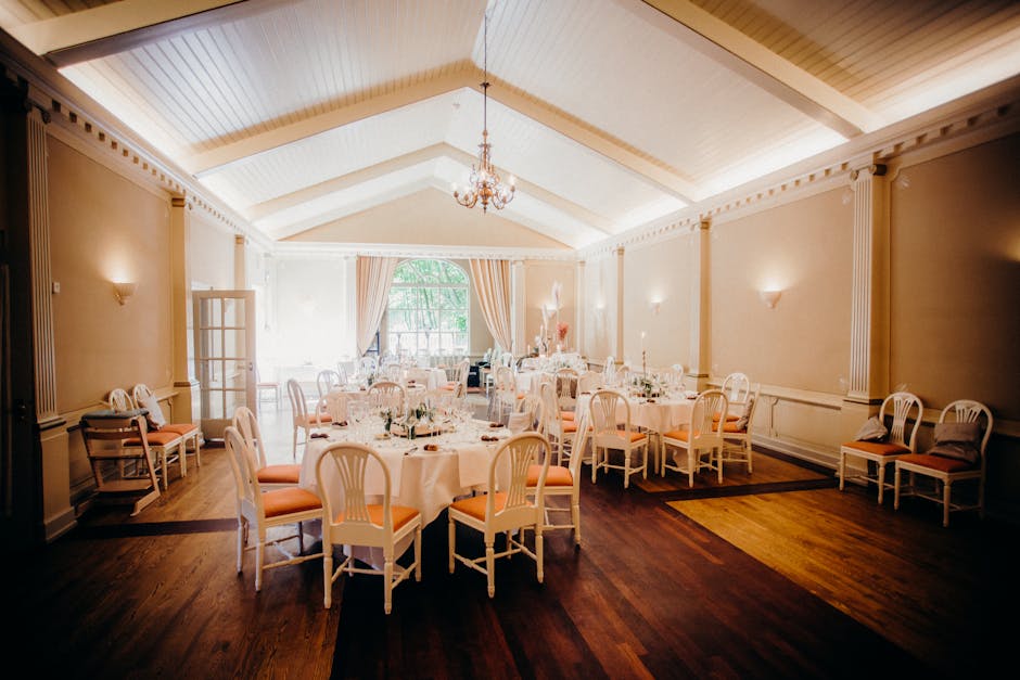 An elegant, bright banquet hall set up for a formal event with round tables covered in white tablecloths and decorated with floral arrangements featuring soft pastel-colored flowers. The arrangement i