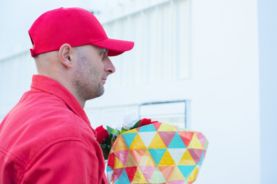 A male delivery person dressed in a red cap and jacket is holding a colorful, geometric-patterned floral bouquet wrapped in pastel paper, with visible red, white, and green flowers. He is standing out