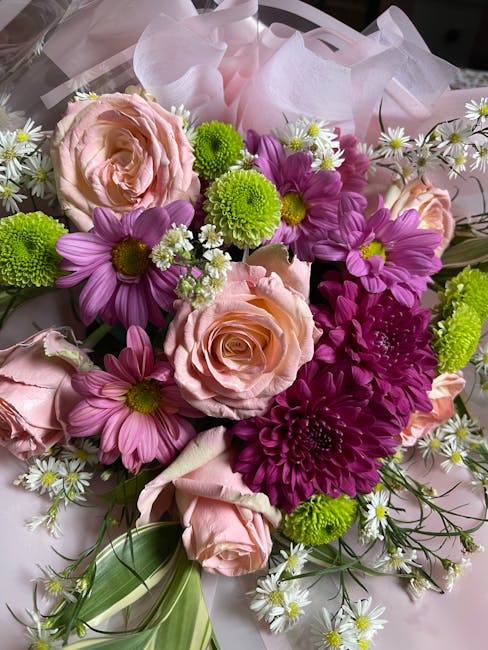 A vibrant bouquet of fresh flowers featuring soft pink roses, purple daisies, and bright green chrysanthemums, delicately arranged with small white daisies and lush green foliage. The flowers are wrap