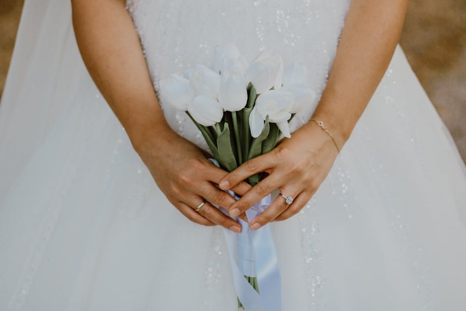 A bride holding a bouquet of white calla lilies with long green stems, wrapped in a white satin ribbon. She is wearing a white wedding dress with delicate pearl embellishments and has her hands gently