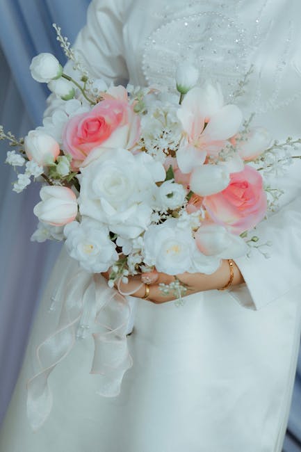 A bride, dressed in a white wedding gown with delicate embroidery details on the bodice, is holding a beautifully arranged bridal bouquet featuring soft pink and white roses, white tulips, and delicat