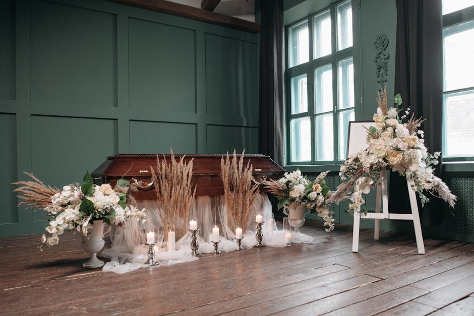 A floral memorial tribute featuring two large cream and white flower arrangements, including roses, orchids, and hydrangeas, set on white vases placed on the floor, with tall beige pampas grass accent