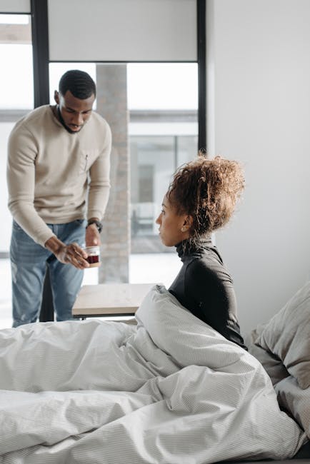 The image depicts a scene in a modern bedroom with natural daylight streaming through large windows. A man in a beige sweater and light blue jeans is standing beside a bed, holding a small jar or cont